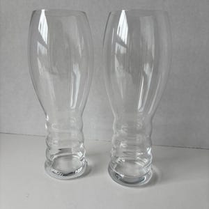 May include: Two clear, tulip-shaped beer glasses. The glasses have a wide, rounded bowl that tapers to a narrow stem with a ridged design. The bases are flat and circular. The glasses are empty and reflect the light.