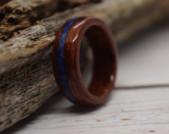 May include: A wooden ring with a polished finish. The ring is made of dark brown wood and features a thin inlay of blue stone. The ring is displayed on a piece of weathered wood.