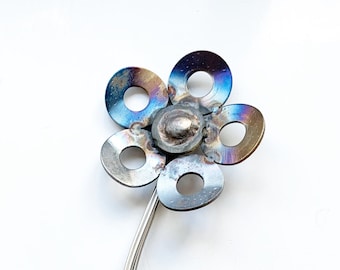May include: A handcrafted metal flower sculpture with a stem and leaves, set in a cylindrical pot. The flower petals are made from circular metal pieces with a blue and purple hue. The text "METAL LAINE ARTISTRY" is visible.