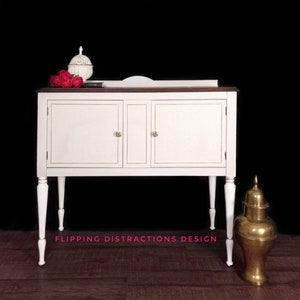 May include: White painted wooden sideboard with two doors and gold knobs. The sideboard has a dark wood top and sits on four turned legs. The sideboard is on a dark wood floor. "Flipping Distractions Design" is written on the floor in front of the sideboard.