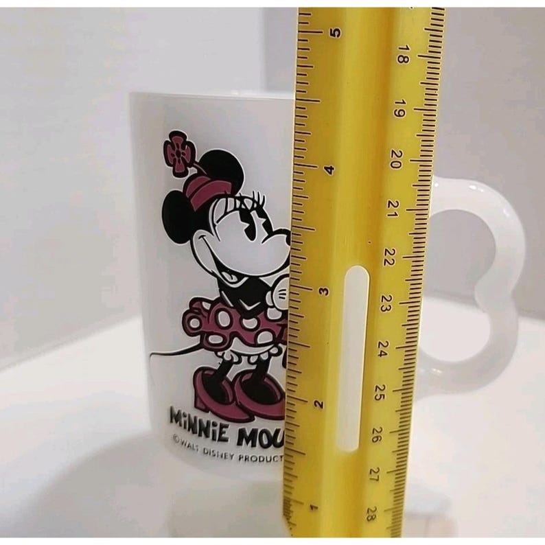 Puede incluir: Taza de cer&aacute;mica blanca con Minnie Mouse con un vestido de lunares rojos y blancos y un lazo. La taza tiene un asa en forma de coraz&oacute;n. La taza mide aproximadamente 10 cm de alto. El texto "MINNIE MOUSE" y "&copy; WALT DISNEY PRODUCT" est&aacute;n impresos.