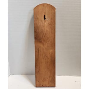 May include: A rectangular wooden plaque with a rounded top and a keyhole for hanging. The wood grain is visible, and the plaque is a warm brown color.
