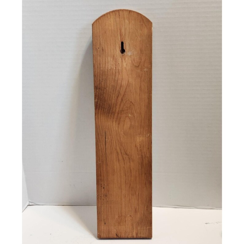 May include: A rectangular wooden plaque with a rounded top and a keyhole for hanging. The wood grain is visible, and the plaque is a warm brown color.