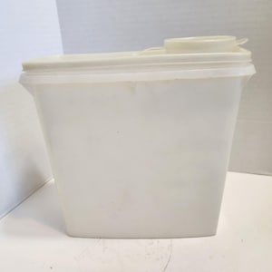 May include: A rectangular, off-white plastic storage container with a slightly tapered base and a flat, removable lid. The lid features a small, circular, raised pouring spout with a separate, smaller cap attached to the top surface.