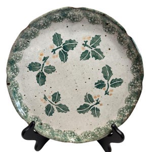 May include: A round ceramic plate with a speckled off-white surface featuring a green holly leaf and berry pattern arranged in a circle. The plate has a scalloped edge with a textured green and white mottled glaze border.