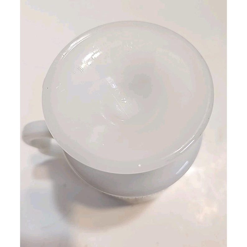 Puede incluir: Vista superior de una taza de cer&aacute;mica blanca con asa. La taza tiene forma redondeada y una superficie lisa y brillante. El interior de la taza tambi&eacute;n es blanco. La taza est&aacute; sobre una superficie blanca.