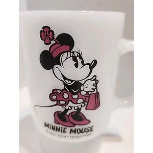 Puede incluir: Taza de cer&aacute;mica blanca con un dise&ntilde;o de Minnie Mouse. El personaje de dibujos animados est&aacute; representado con un atuendo rosa y negro, sosteniendo un bolso rosa. La taza tiene un asa y las palabras "MINNIE MOUSE" est&aacute;n impresas en la parte inferior.
