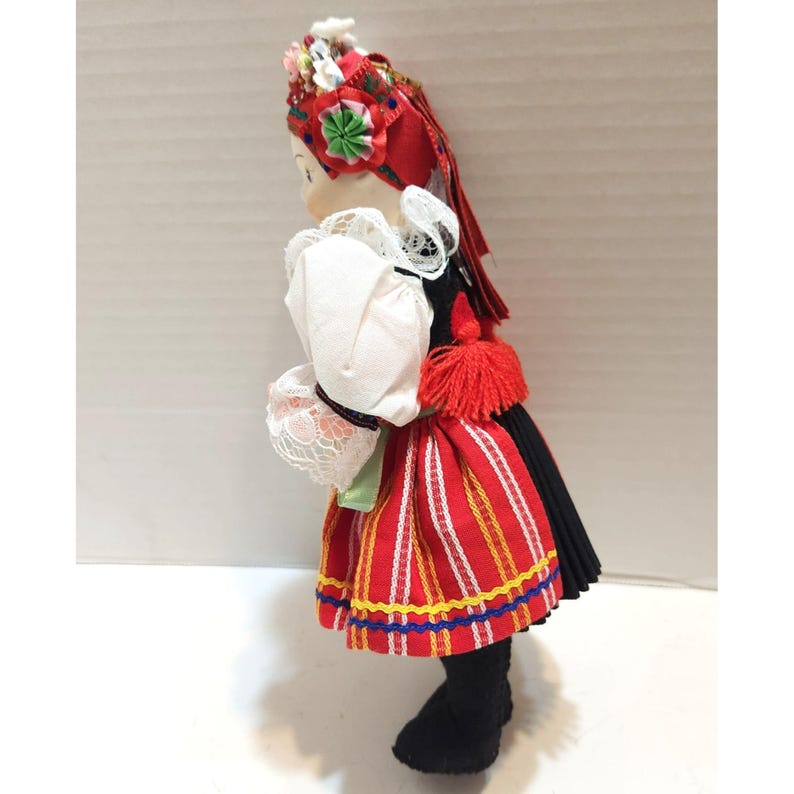 May include: A Polish folk doll in traditional attire. The doll wears a red and white striped skirt, a white blouse with lace trim, and a black bodice. A red headdress with floral accents and a red tassel complete the outfit. The doll's black boots are visible.