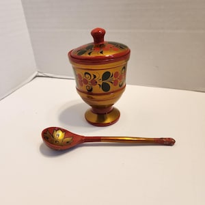 Peut inclure: Un sucrier en bois peint à la main avec un couvercle et une cuillère assortie. Le bol et la cuillère sont peints avec des motifs floraux dorés, rouges et noirs. Le bol a une base en piédestal et une forme arrondie.