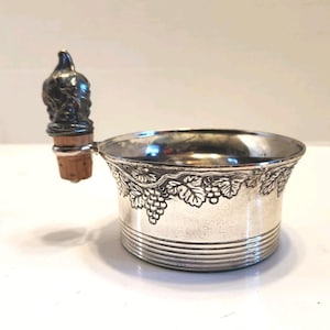 May include: Silver wine bottle coaster with a decorative grape and leaf design around the rim. The coaster has a series of horizontal lines near the base. A wine bottle stopper with a detailed figure is attached to the side.