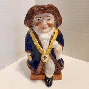 Puede incluir: Figura de cerámica de un hombre con abrigo azul, peluca marrón y cadena dorada. La figura sostiene un pergamino y tiene una expresión severa. La base es marrón y la figura mide aproximadamente 15 cm de altura.