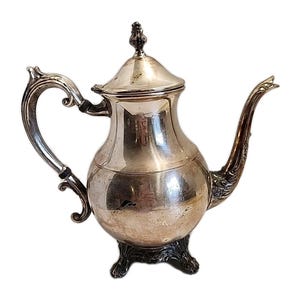 Puede incluir: Una tetera plateada con tapa, asa curva y boquilla delgada. La tetera tiene un cuerpo bulboso y se asienta sobre una base decorativa. El asa y la boquilla tienen detalles ornamentados.