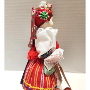 May include: A porcelain doll in traditional Polish folk attire. The doll wears a red and white striped skirt, a white blouse with lace trim, and a red headdress adorned with flowers and a green rosette. The doll's sleeves have a floral embroidered cuff.