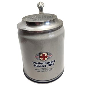 Op de afbeelding: Een lichtgrijze keramische bierpul met een metalen deksel voorzien van een schelpvormige duimsteun. De voorzijde toont het Weltenburger Kloster Bier logo met een rood kruis, de tekst Seit 1050 en de zin Aus der ältesten Klosterbrauerei der Welt.
