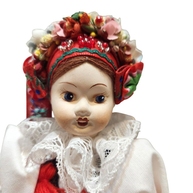 May include: A porcelain doll with blue eyes and a detailed red headdress adorned with flowers and ribbons. The doll wears a white dress with a lace collar and a red tassel. This collectible doll showcases traditional folk attire.