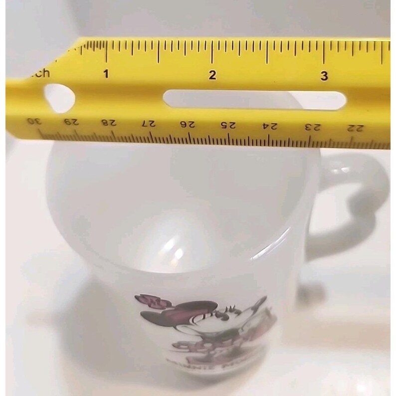 Puede incluir: Taza de cer&aacute;mica blanca con un gr&aacute;fico de Minnie Mouse. La taza tiene aproximadamente 6,35 cm de di&aacute;metro, medido con una regla amarilla. El asa es blanca y curvada. El gr&aacute;fico de Minnie Mouse incluye colores rojo, negro y rosa.