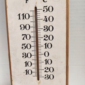 May include: A vintage thermometer with Fahrenheit and Celsius scales. The wooden frame surrounds a cream-colored face with black numerals. The thermometer reads approximately 10 degrees Fahrenheit, equivalent to -12 degrees Celsius.