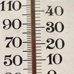 May include: Close-up of a vintage thermometer with black numbers against a cream-colored background. The temperature scale ranges from 0 to 110 degrees Fahrenheit. A red liquid column indicates the current temperature. The thermometer is used to measure temperature.
