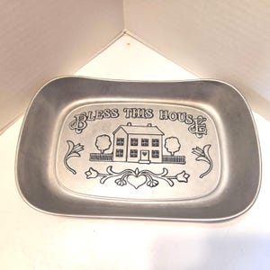 Op de afbeelding: Een rechthoekige, tin-kleurige schaal met afgeronde randen. De schaal heeft een gegraveerd ontwerp met de woorden "BLESS THIS HOUSE" boven een huisillustratie, bomen en bloemaccenten. Een decoratief stuk voor in huis.