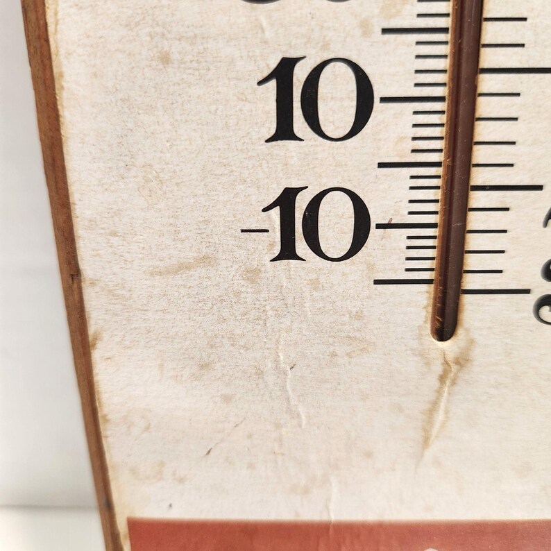 May include: Close-up of a vintage thermometer with a wooden frame. The thermometer displays the numbers 10 and -10 in black against a cream-colored background. A brown temperature gauge is visible on the right side, with temperature markings.