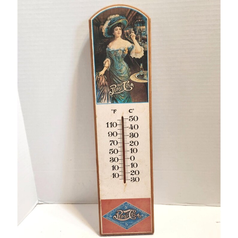 May include: Vintage Pepsi Cola thermometer with a wooden frame. The top features an illustration of a woman in a teal dress and hat. The thermometer displays Fahrenheit and Celsius scales, with a range from -30 to 110 degrees Fahrenheit.
