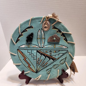 Peut inclure: Une décoration murale circulaire en daim turquoise ornée de motifs géométriques peints à la main en noir, blanc et orange. L'objet est lacé avec un cordon en cuir beige sur les bords et décoré d'une pointe de flèche en obsidienne noire.