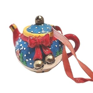 May include: A ceramic Christmas ornament shaped like a teapot. The teapot is painted with a blue background and white polka dots, a red bow, and gold bells. The handle and spout are red, and the lid is yellow.