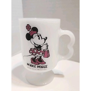 Puede incluir: Taza blanca con pedestal con Minnie Mouse. La taza tiene un asa con dise&ntilde;o de coraz&oacute;n. Minnie Mouse est&aacute; representada con un vestido rosa de lunares, sosteniendo un bolso rosa. La taza est&aacute; marcada con "MINNIE MOUSE" y "WALT DISNEY PRODUCTIONS".