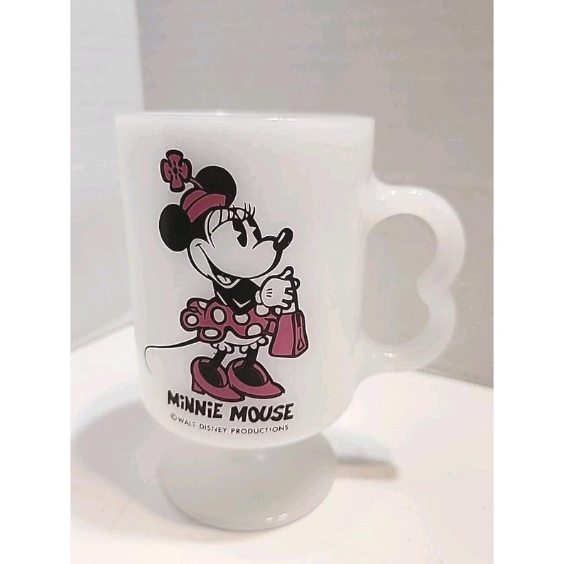 Puede incluir: Taza blanca con pedestal con Minnie Mouse. La taza tiene un asa con dise&ntilde;o de coraz&oacute;n. Minnie Mouse est&aacute; representada con un vestido rosa de lunares, sosteniendo un bolso rosa. La taza est&aacute; marcada con "MINNIE MOUSE" y "WALT DISNEY PRODUCTIONS".
