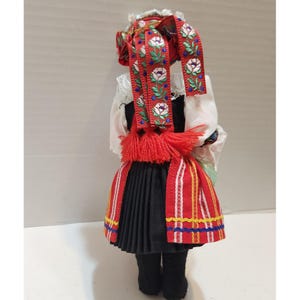 May include: A traditional Polish doll, viewed from the back, wearing a detailed folk costume. The doll features a red and white headpiece with floral embroidery, a black pleated skirt, and a red and white striped apron. The doll's outfit includes a white blouse and black boots.