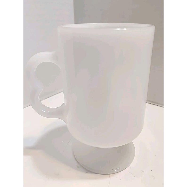 Puede incluir: Una taza de vidrio blanco opaco con un asa en forma de coraz&oacute;n. La taza tiene una base de pedestal y un cuerpo cil&iacute;ndrico. El asa es un lazo con forma de coraz&oacute;n. La taza probablemente se usa para bebidas calientes.