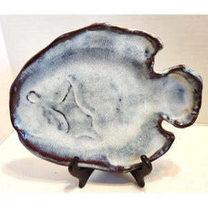 Puede incluir: Plato de cerámica con forma de pez, con interior moteado blanco y azul y borde marrón oscuro. El plato se exhibe sobre un soporte negro. El diseño del pez es visible en el interior del plato.