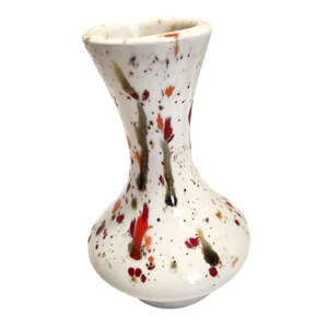 May include: A small ceramic vase with a white base featuring a speckled pattern of red, orange, and dark brown paint splatters. The vase has a rounded body that tapers into a narrow neck and flares slightly at the top opening, with a smooth finish.