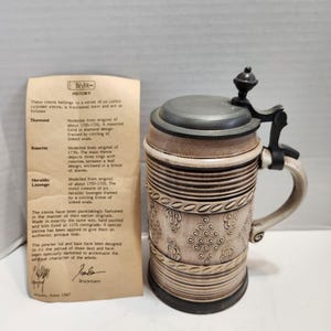 May include: A beige ceramic beer stein with a dark grey pewter lid and handle. The stein features a textured relief pattern of diamonds and rosettes. Beside the stein is a tan paper document titled Beyer History with printed text.