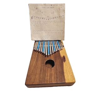 May include: A wooden Treble Kalimba musical instrument with metal tines and a sound hole. The instrument has a dark wood finish and blue accents. A paper card with musical notes and instructions is placed on top of the instrument.