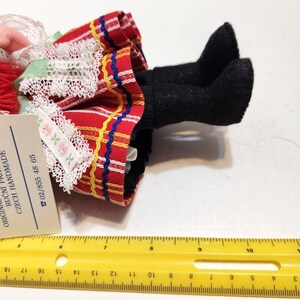 May include: A handmade doll with black felt legs and a red skirt with yellow and blue stripes. The skirt has white lace trim and a tag with text. A yellow ruler is in the foreground.