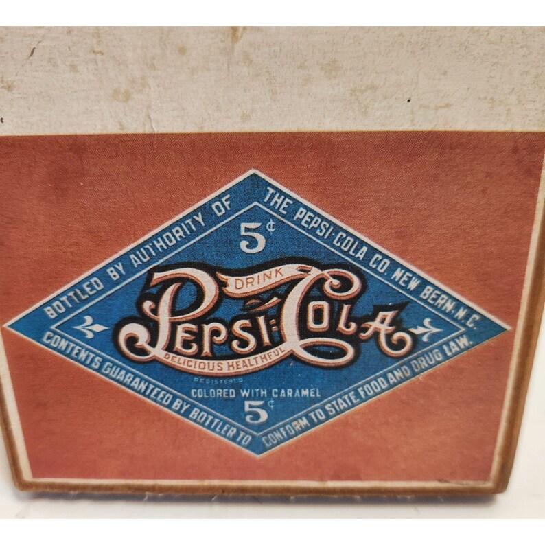 May include: Vintage Pepsi-Cola advertisement featuring a diamond-shaped logo with the Pepsi-Cola name in a stylized script. The logo is set against a red background, with text indicating the drink is "Delicious, Healthful" and costs 5 cents.