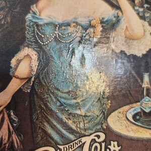 May include: Vintage advertisement featuring a woman in a blue dress with pearl accents. The image includes a bottle of cola on a small table. The text "Drink Cola" is visible at the bottom of the image.
