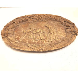 May include: Oval, light brown, carved wooden tray with a relief depicting figures harvesting wheat. The tray's border features wheat stalks and leaves. The figures are dressed in long robes, and the tray has a distressed finish.
