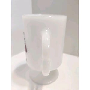 Puede incluir: Taza de caf&eacute; blanca con pedestal y asa curva. La taza tiene un cuerpo ancho y redondeado y una base peque&ntilde;a y circular. Una imagen parcial de un dise&ntilde;o es visible en el lateral de la taza.