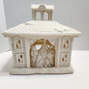 May include: A cream-colored ceramic house figurine featuring a shingled roof with snow accents and a cupola. The open front reveals a relief scene of two adults and a child decorating a Christmas tree, with arched windows on the side walls.
