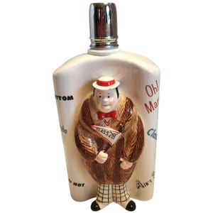 Może przedstawiać: Wazon ceramiczny w stylu vintage ze srebrnym korkiem. Wazon przedstawia płaskorzeźbę mężczyzny w brązowym futrze, czerwonej muszce i białym kapeluszu z czerwoną opaską. Mężczyzna trzyma znak z napisem "CLASS 28". Wazon ma tekst po bokach.