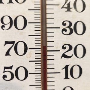 May include: Close-up of a vintage thermometer with black numbers and markings against a cream-colored background. The red liquid indicates a temperature of approximately 72 degrees Fahrenheit.