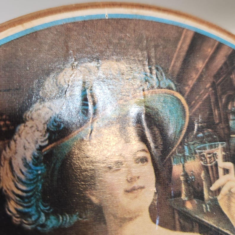 May include: A vintage, round wooden tray with a painted image of a woman in a teal hat holding a glass of dark beer. The image has a classic, antique aesthetic, with a brown and blue border.