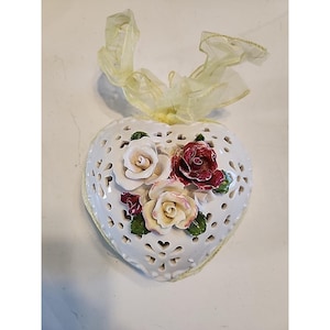 May include: White ceramic heart-shaped wall decor with a delicate openwork design. The heart is adorned with three-dimensional roses in shades of white, cream, and burgundy, with green leaves. A yellow ribbon is attached at the top.