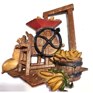 May include: A rustic, three-dimensional wall decoration depicting a farm scene. The piece features a red hopper, a black wheel, and a wooden structure with a barrel and several ears of corn. The color palette includes shades of brown, yellow, and green.