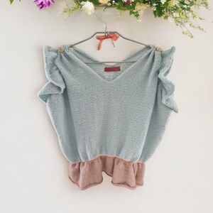 May include: A light blue knitted top with a ruffled hem and ruffled sleeves. The top has a V-neck and gold embellishments on the shoulders.