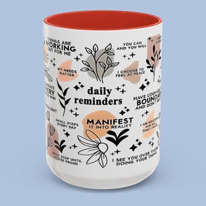 Puede incluir: Una taza de cer&aacute;mica blanca con interior rojo. La taza est&aacute; decorada con frases inspiradoras y dise&ntilde;os florales en negro, melocot&oacute;n y gris. Las palabras "daily reminders" son prominentes.