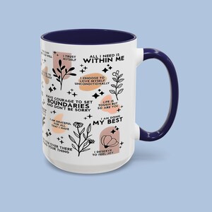 Puede incluir: Taza de cer&aacute;mica blanca con interior y asa azul marino. La taza presenta frases inspiradoras e ilustraciones bot&aacute;nicas en negro y melocot&oacute;n. El texto incluye "Todo lo que necesito est&aacute; dentro de m&iacute;".