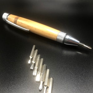 May include: A wooden pen-style screwdriver with a silver clip and tip, and a set of silver screwdriver bits. The screwdriver has a light brown wooden barrel and a silver tip. The bits are arranged in a row.
